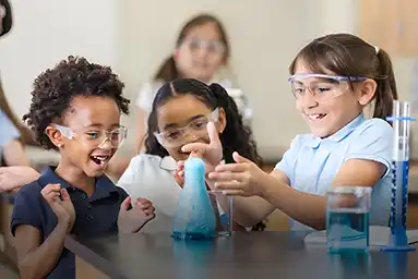 Three smiling children with safety glasses laughing around a science beaker